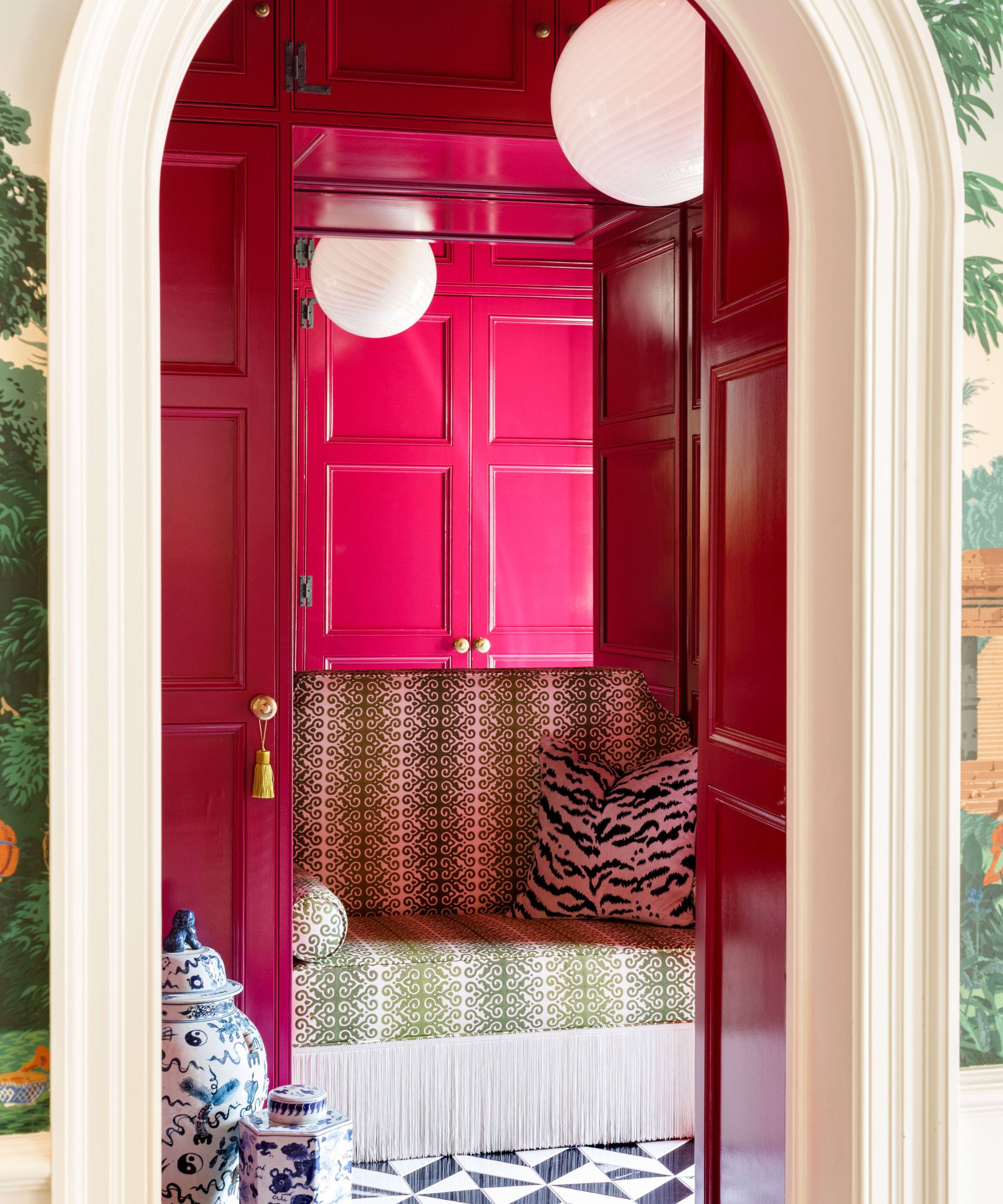 Cloakroom with raspberry pink painted cabinetry and walls, black and white geometric floor tiles, a built-in bench with fringe and green striped fabric, a tiger print cushion and antique ginger jars