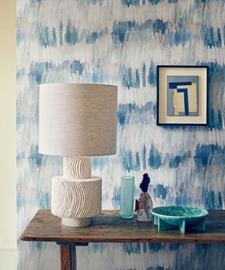 Corner of a living room with blue and gray brushed wallpaper design, dark wooden shelf, cream lampshade with textured base, ceramic and glass decorative ornaments on shelf