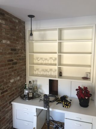 A home bar in an alcove with a white desk, white shelving, and an exposed brick wall