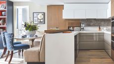 A bright kitchen with white cabinets and wooden flooring. To the left is a comfortable seating/ dinign area with a beige sofa chair, table, and blue patterned seats with wooden legs. To the far left is a blue bookshelf.