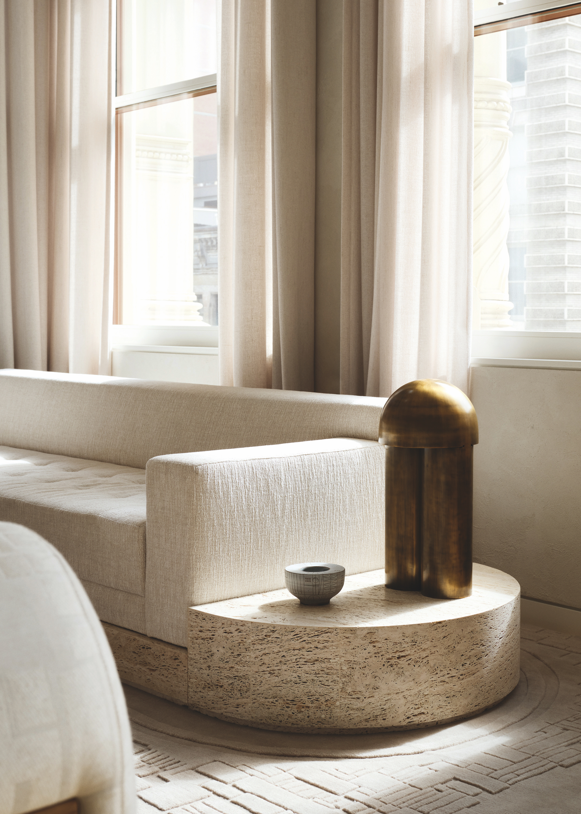 A modern, cream and beige living space with a cream built in sofa in from of two large windows, with a stone side table connected to the sofa, with a geometric cream rug underneath.
