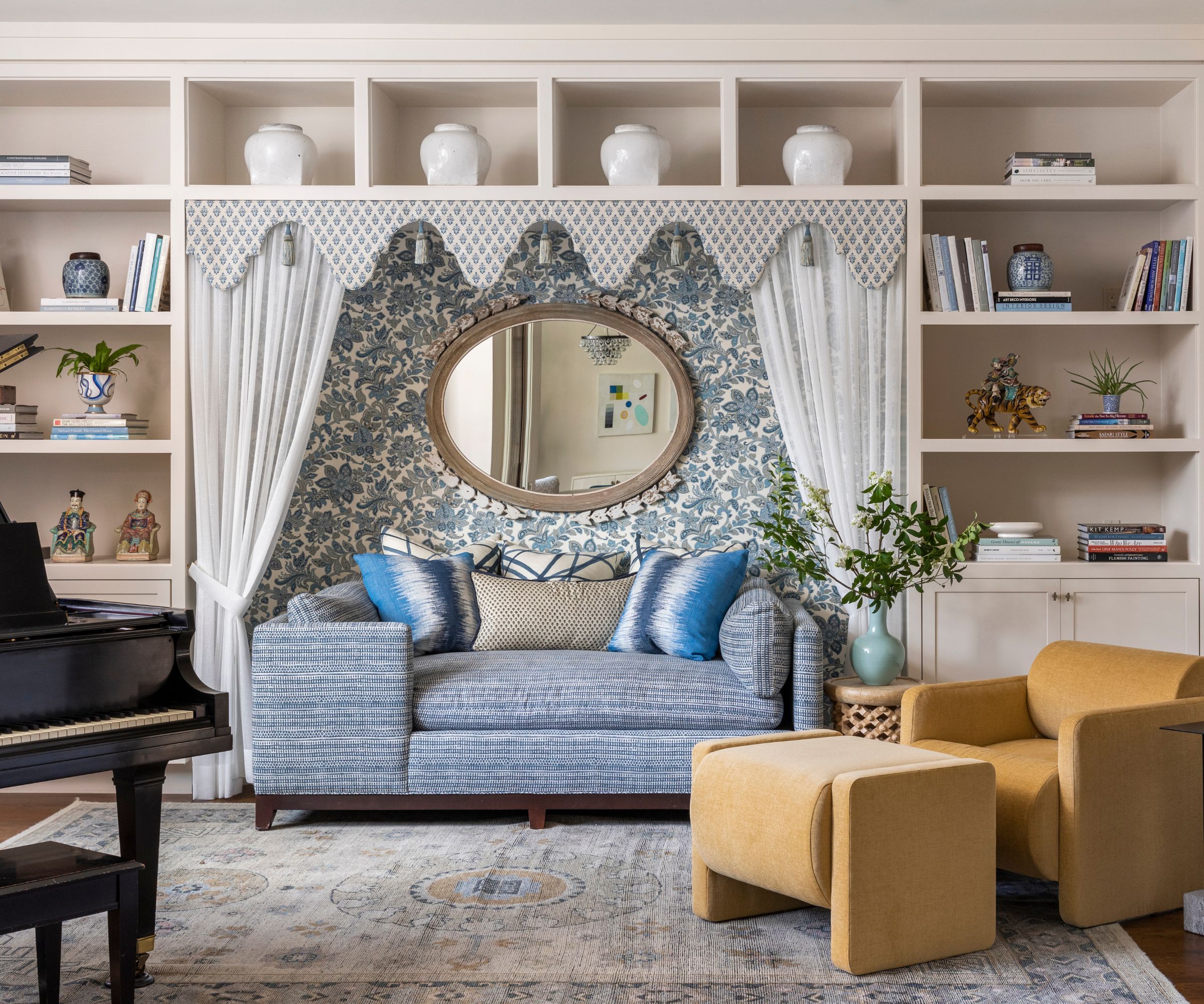 A coastal living room with a wall of white built-ins with blue and white decorative objects, a blue floral wallpaper feature wall in the middle with white curtains, a blue sofa with blue cushions, and an ochre accent chair.