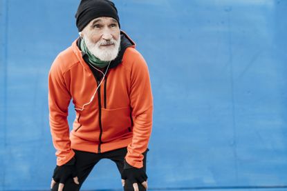 An older retired man rests in front of a blue wall after jogging. He is smiling and resting his hands on his knees. He wears a winter cap and bright orange hoodie.