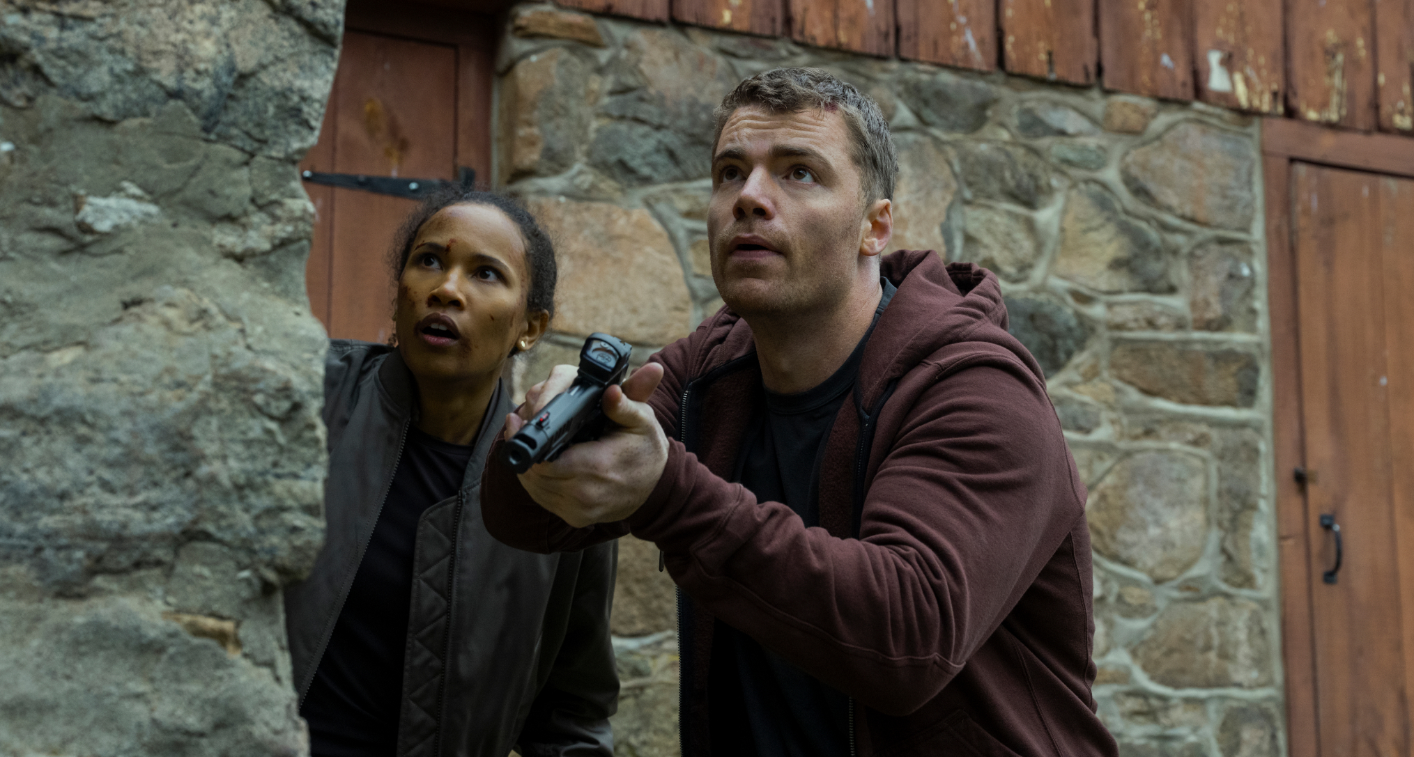 Fola Evans-Akingbola as chelsea and Gabriel Basso as peter sutherland pointing a gun and hiding behind a brick wall while investigating in the night agent season 3