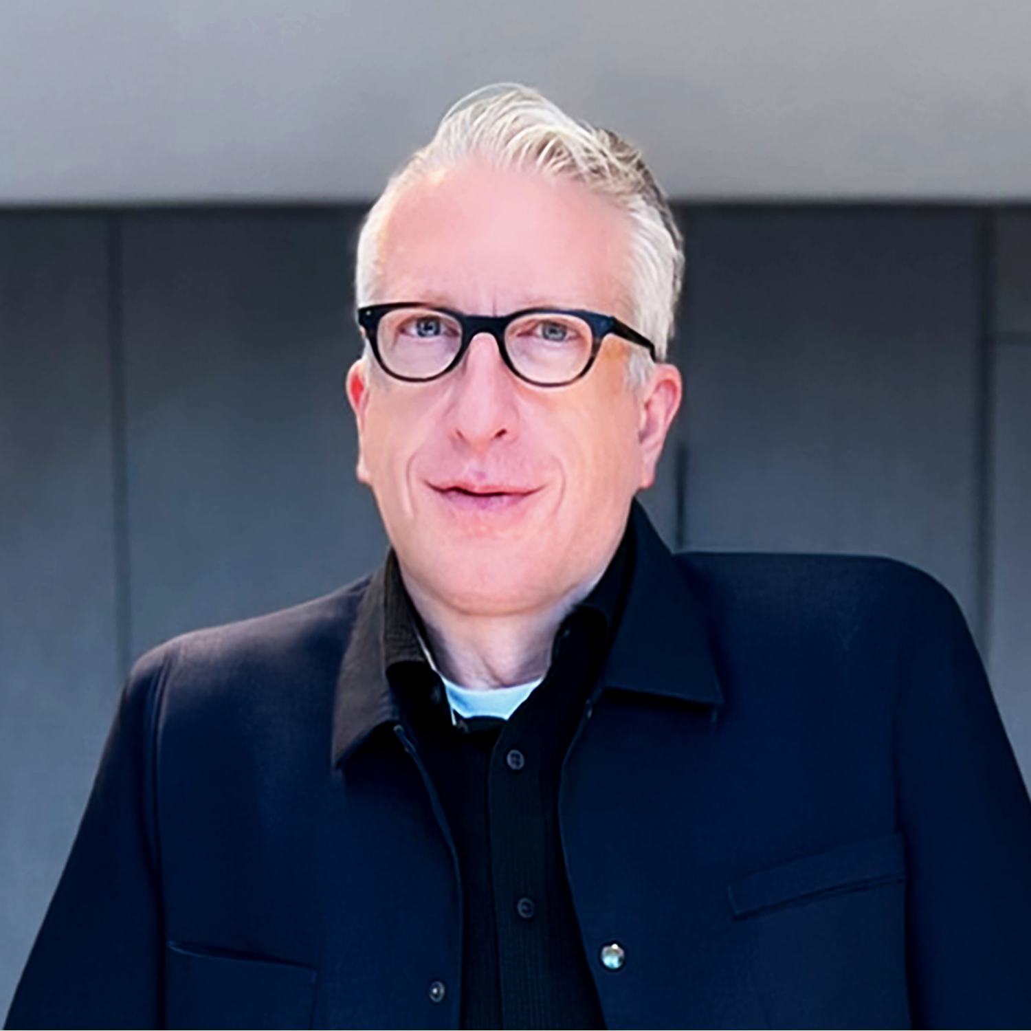 A headshot of a man with white hair, black glasses, a dark blue coat and a black shirt