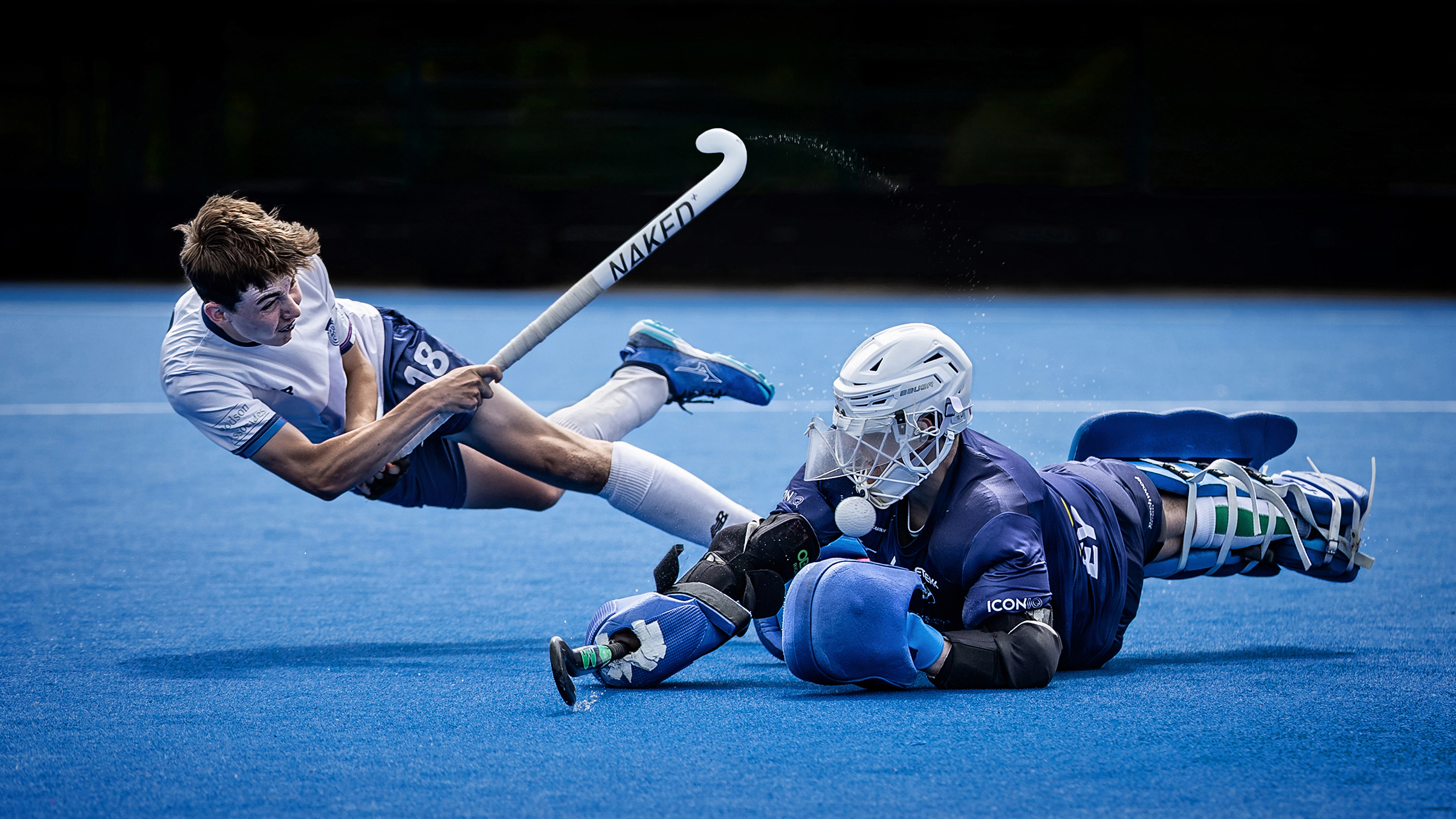 A hockey player lunges toward the goal while a goalkeeper dives to block a shot, set against a vibrant blue turf field
