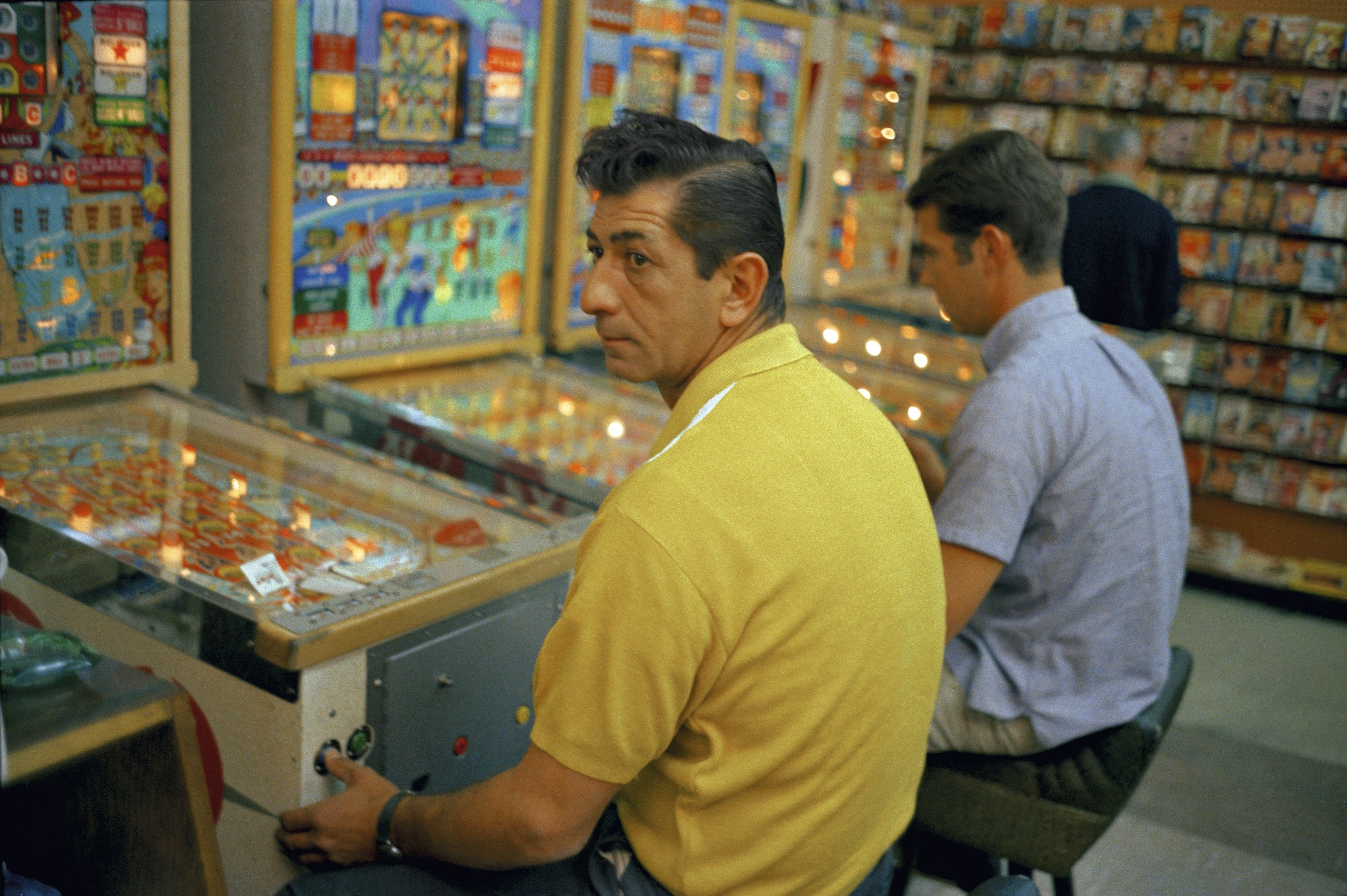 A color photograph of two men playing pinball machines inside an arcade lined with rows of lit-up games.