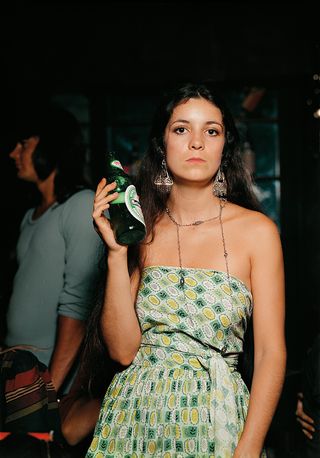 View of a woman with long dark hair holding a bottle of Heineken at a night club. She is wearing earrings, a necklace and a green, white and yellow patterned sleeveless outfit. There is a man wearing a blue long sleeved top in the background