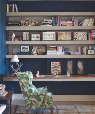 A blue living room wall with white shelves with books and decor on with a green leafy chair in front of it