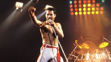 Freddie Mercury topless on stage in white pants with a red belt. He is holding a microphone and mid note