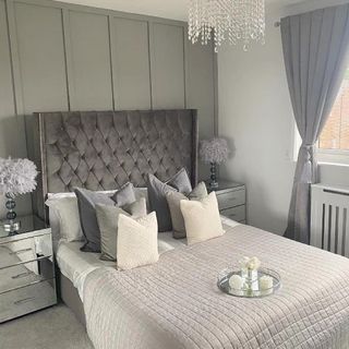 an elegant bedroom in multiple tones of grey, large elegant headboard and bed cushions, mirrored bedside cabinets in front of a grey panel wall