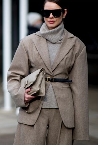 One of the best designer belts is shown in a close street-style photo of a woman wearing a tan blazer over a gray turtleneck with matching trousers, a black Saint Laurent belt, and a tan clutch bag