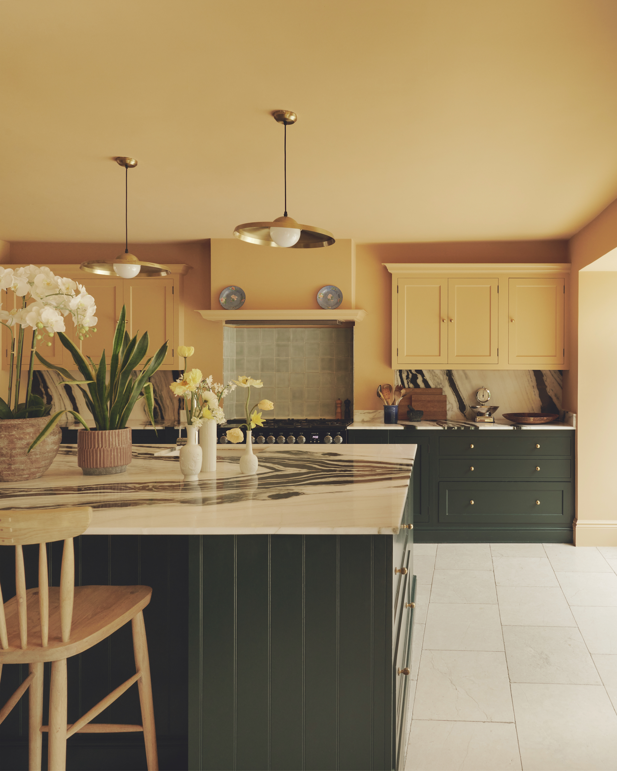 A traditional style kitchen with olive green cupboards, a marble island and work tops and butter yellow cupboards above. There are two pendant lamps above the island and a tiled backsplash in the alcove of the hob.