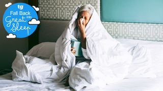 A woman wrapped in a duvet in bed holding a box of blue tissues and blowing her nose as she struggles with the symptoms of a cold
