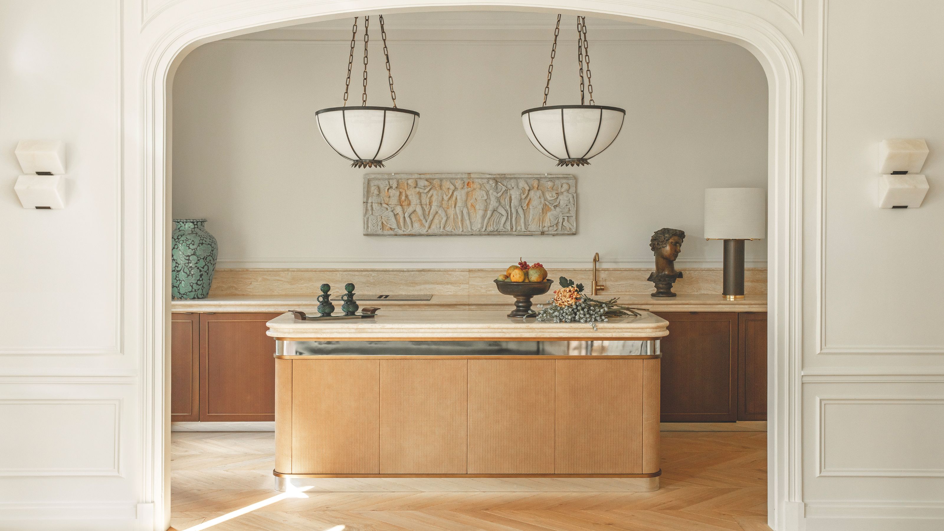 A sleek wooden kitchen viewed through an arched doorway, featuring a light wood island with a stone countertop, marble backsplashes, and two large bowl-shaped pendant lights hanging from chains.
