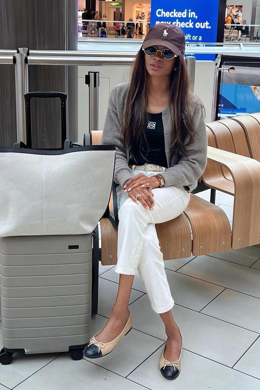 Summer airport outfit is shown in a photo of a French woman sitting in an airport wearing a brown baseball cap with gold wireframe sunglasses, a gray cardigan, a black Loewe tank top, a white belt, white straight leg jeans, tan and black ballet flats, a gray oversized tote bag, and a gray carry-on suitcase