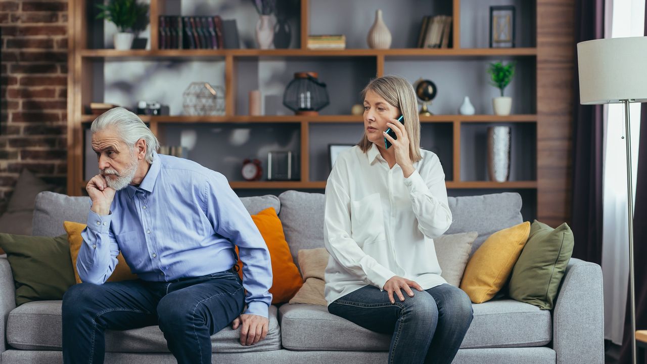 An older couple sit tensely on the sofa, facing away from each other and talking on their own smartphones.