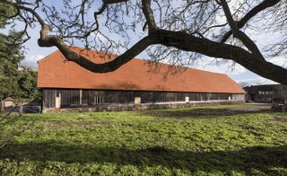 Daytime image of Harmondsworth Barn, red brick roof, metal gate at the left, long patchy grass area to the front, close up of a tree branch dominating the shot, additional builidng to the right in the distance, blue cloudy sky