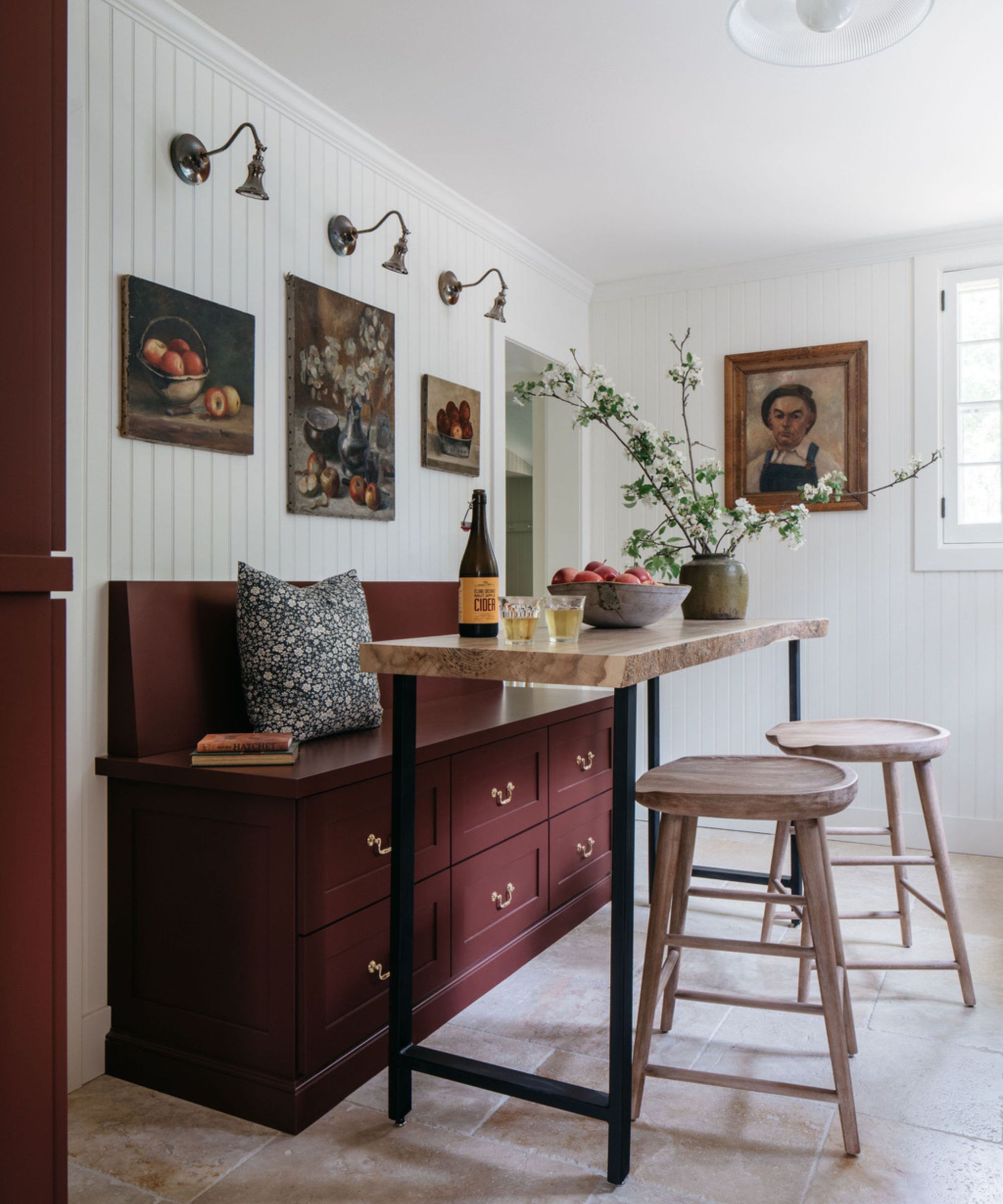 A cottage kitchen eat-in area with a burgundy banquette with two rows of drawer storage and a tall island-style table and two wooden counter stools. On the wall, three vintage artworks are hung and highlighted by brass sconces