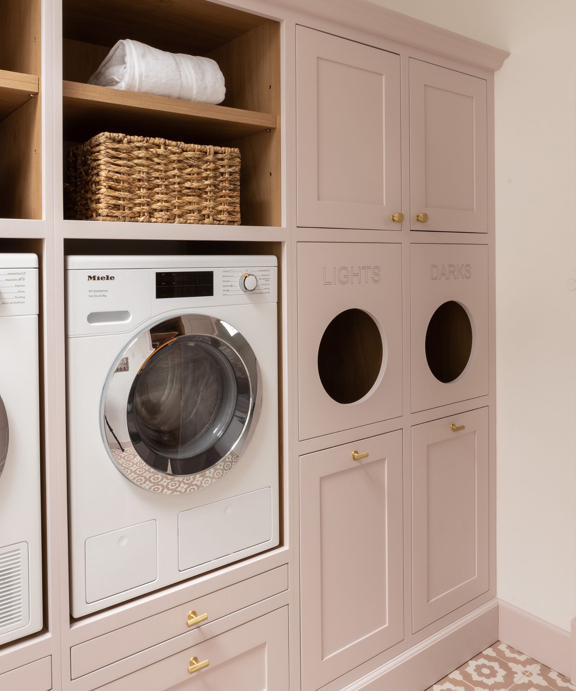 A blush pink fitted utility room with brass handles, a built-in washing machine and a clever laundry sorter cabinet with circular drop holes labelled "Lights" and "Darks".