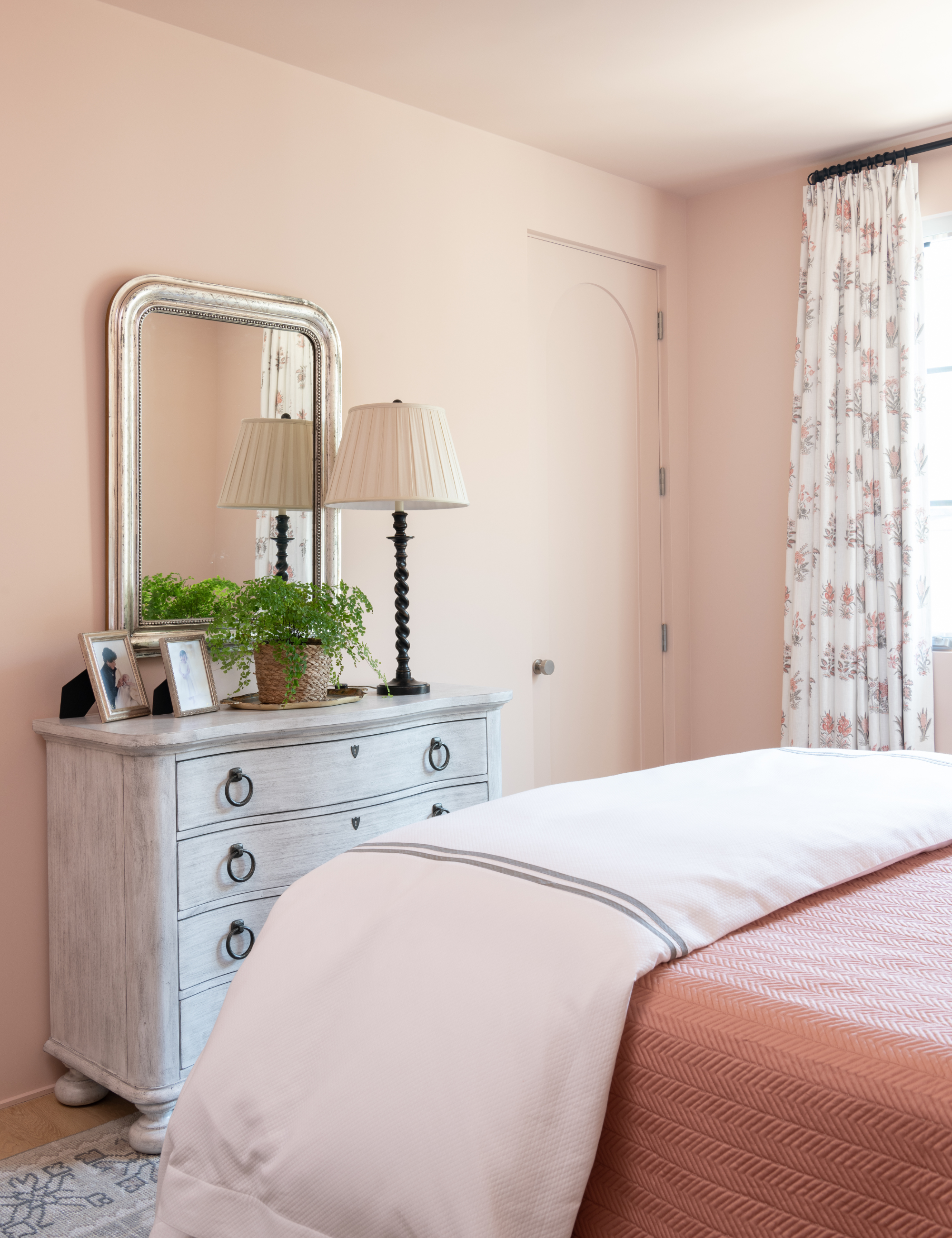 Peachy pink bedroom featuring an antique-style dresser topped with a mantel mirror, leafy potted plant, and linen table lamp.