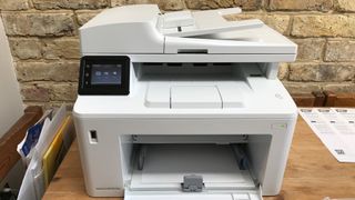 HP LaserJet Pro MFP M227fdw on a wooden desk against a brick wall with documents and print outs either side