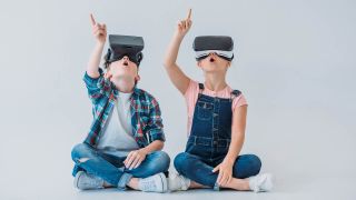 Two kids wearing virtual reality headsets point upward