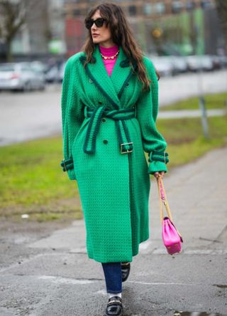 Julie Sergent Ferreri wears black sunglasses, a neon pink turtleneck pullover, a gold pendants necklace, a green embossed wool belted oversized long coat, a neon pink shiny leather with gold nailed / studded handle shoulder bag from Valentino, navy blue denim pants, white socks, black shiny varnished leather with gold logo charms loafers from Chanel, outside Aeron , during the Copenhagen Fashion Week Autumn/Winter 2023 on January 31, 2023 in Copenhagen, Denmark