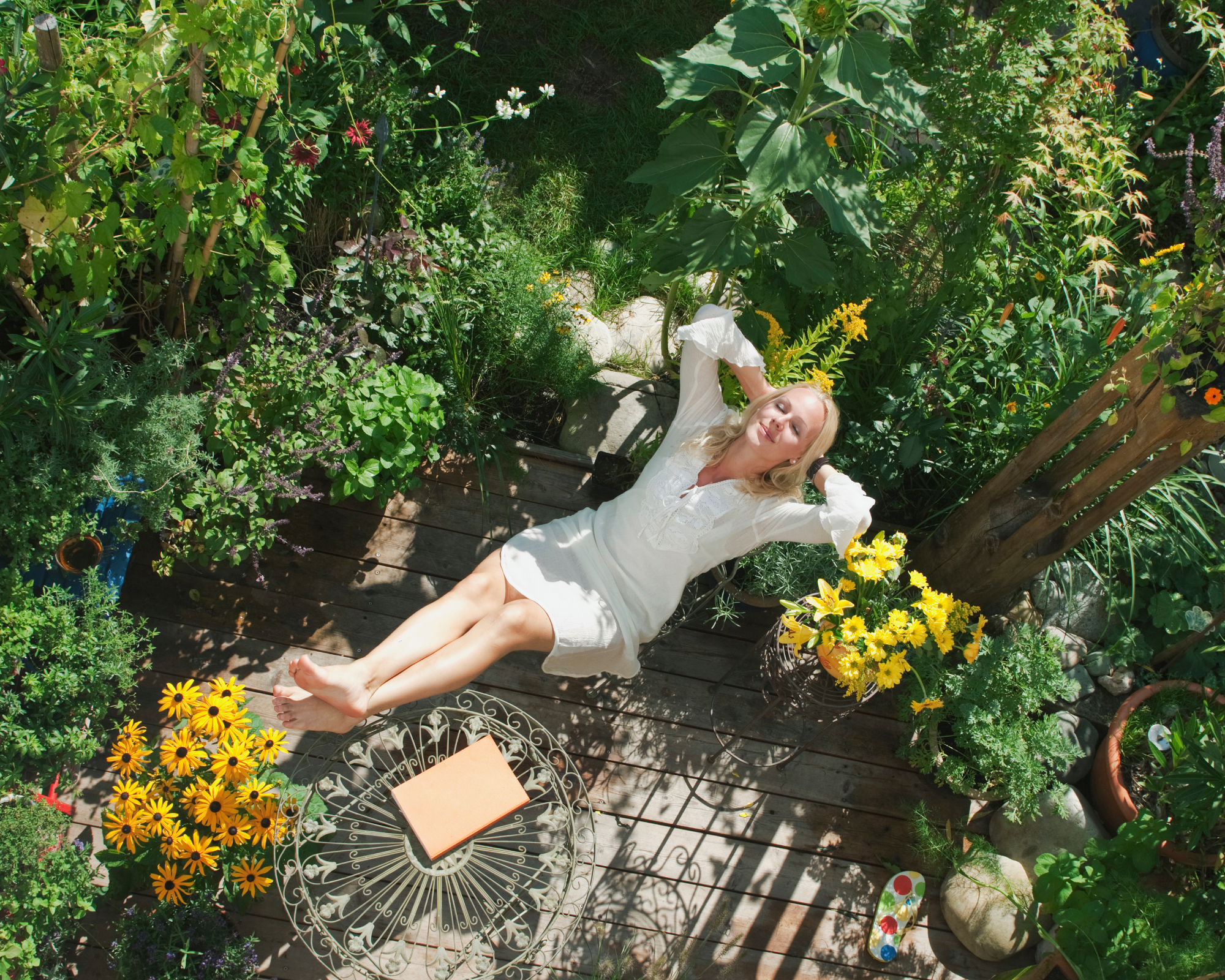 Woman wearing white dress relaxing on a garden deck, surrounded by lush foliage and pots of yellow flowers
