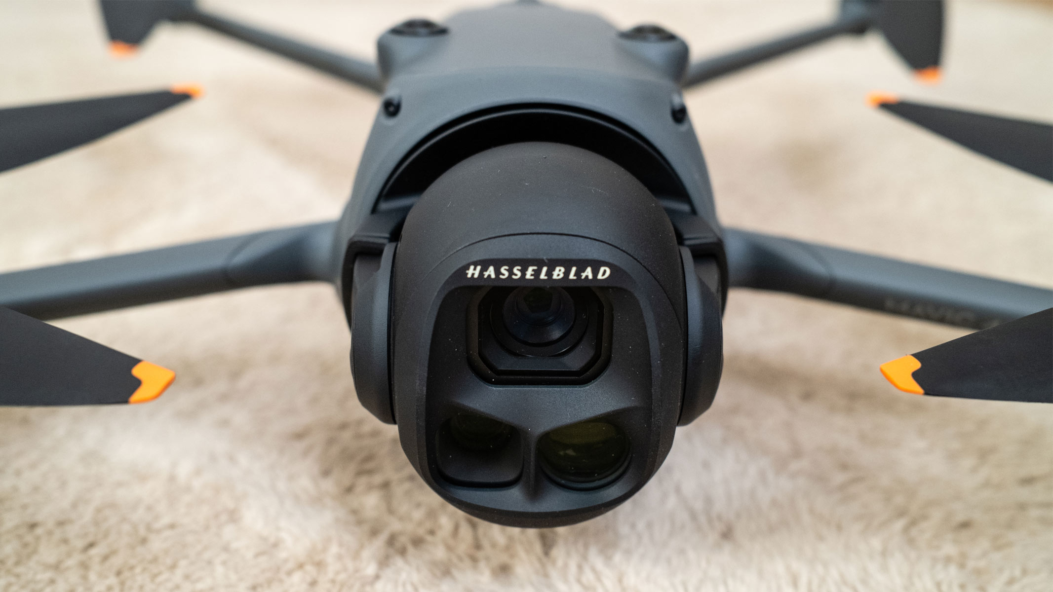 A dark grey drone with four propellers features a black camera on its front, you can see the camera close up as the drone sits on a beige carpet.
