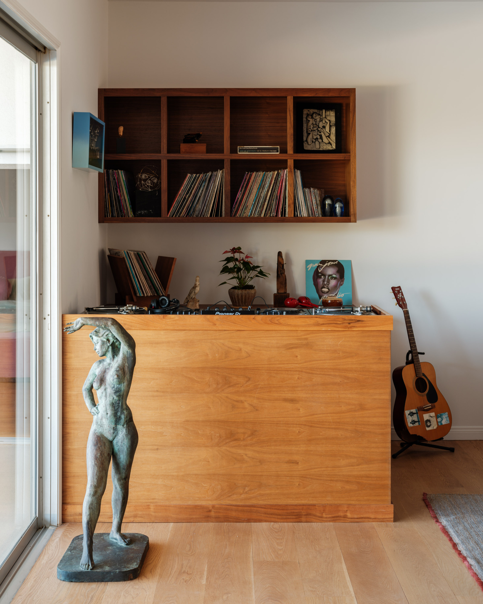 Image of a vinyl bar in the corner of a white living room. There is a large wooden DJ bar with a statue in front of it and a floating shelf on the wall behind it. There is a guitar in the corner as well.