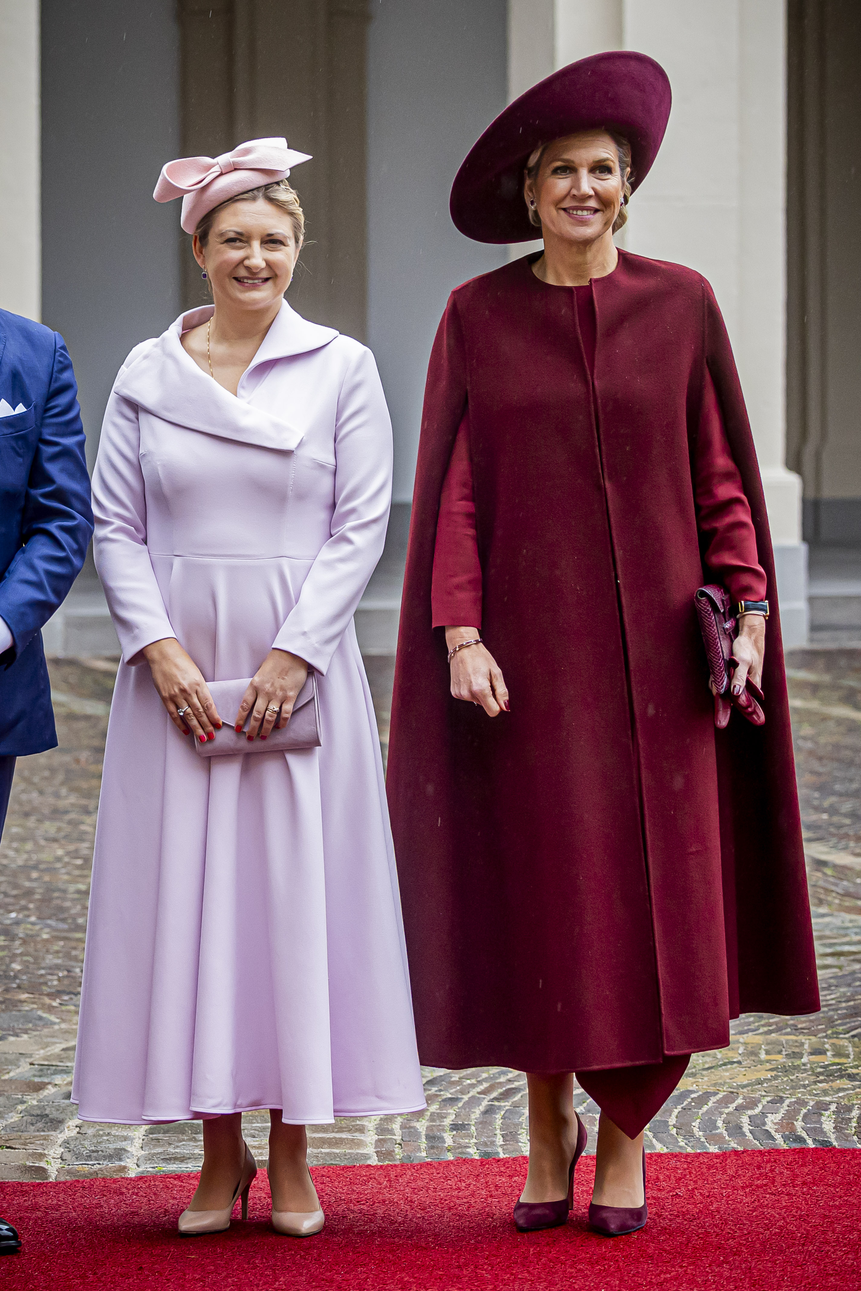 Queen Maxima in a red dress and maroon cape standing next to Grand Duchess Stephanie in a lavender coat