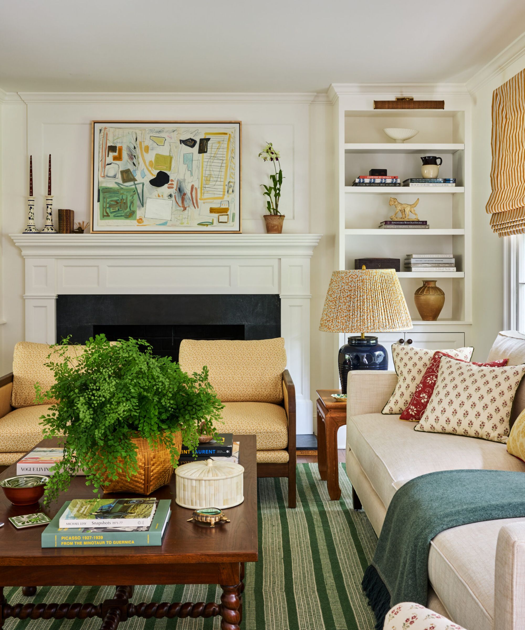 A cozy living room with warm white walls and traditional architectural details. A fireplace with abstract art above, a green striped rug, a wooden coffee table, two pale yellow armchairs, and a cream sofa.