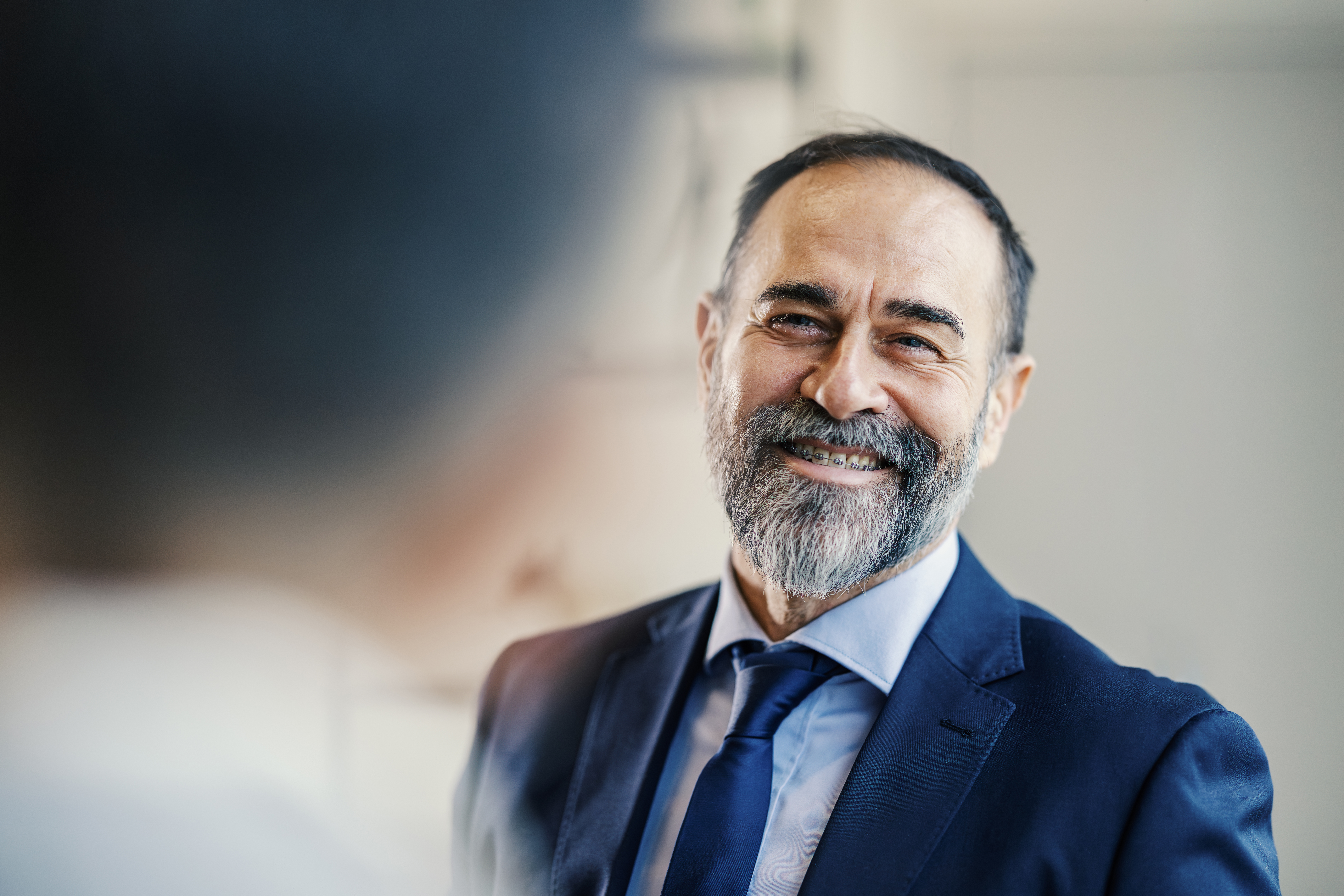 Senior adult businessman confidently smiling, wearing a blue suit and tie, showcasing a friendly and approachable demeanor in a professional office setting, embodying leadership and success