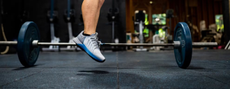 A person jumping next to a barbell in the gym wearing SportsShoes footwear