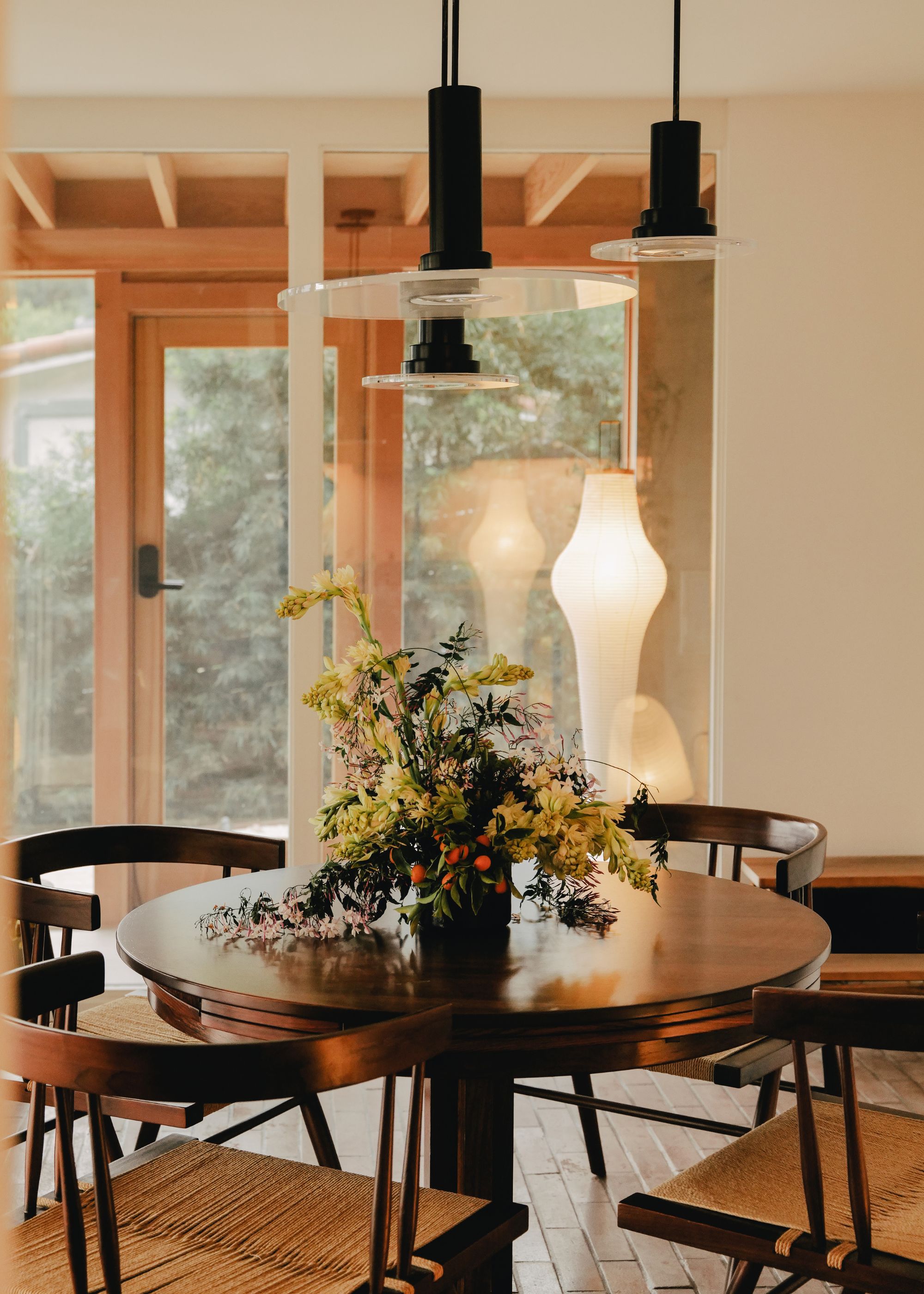 A dining room with overhead pendants, a standing rice paper lamp, a round table with matching chairs, and an overflowing floral arrangement