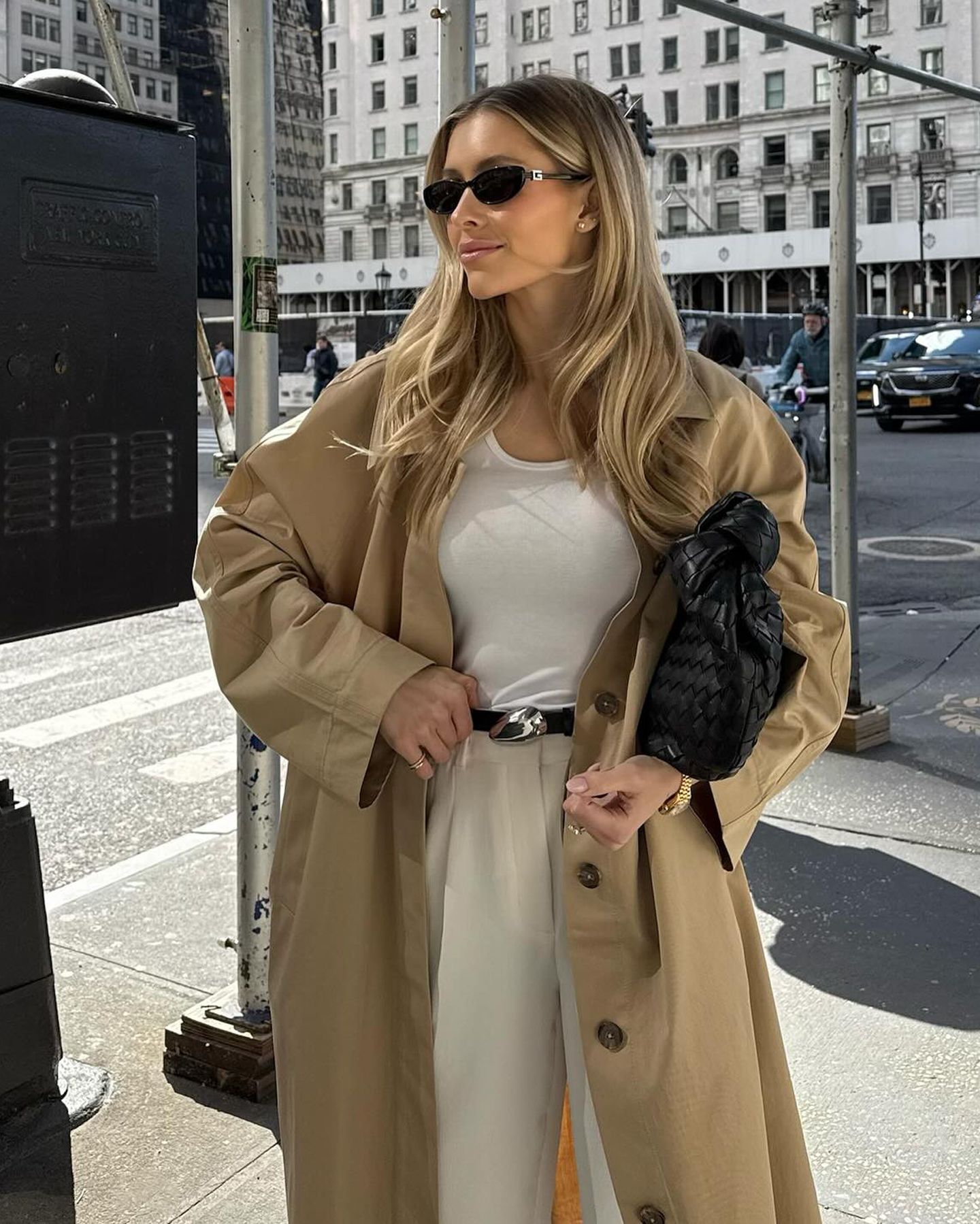 Fashion influencer Fredricka Galgano in New York City in black oval sunglasses, black leather woven bag, white top, black belt with silver sculptural buckle, beige pants, and an oversize trench coat.