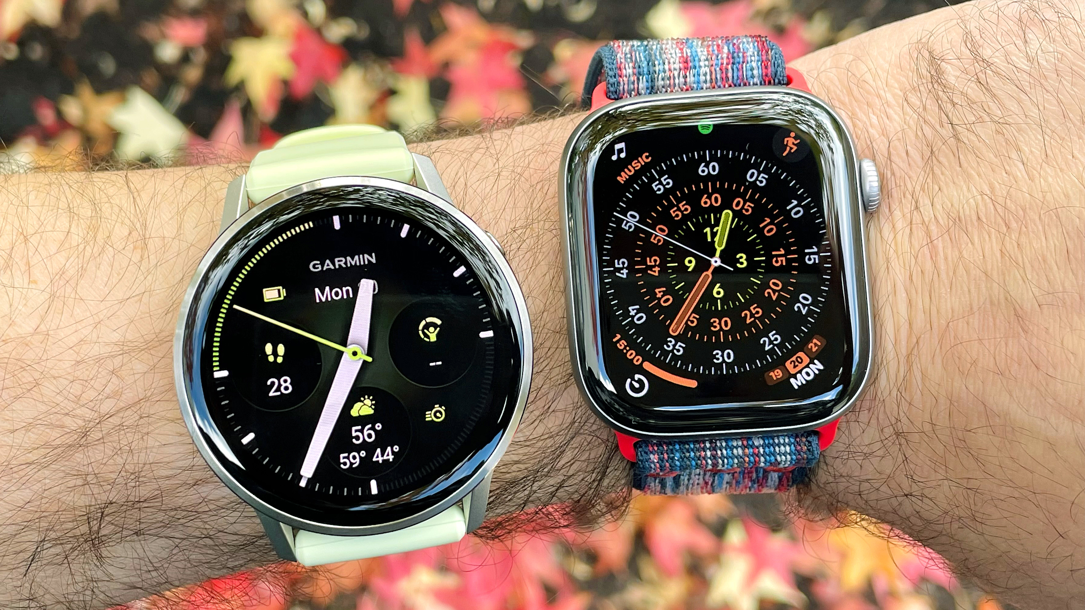 Close-up of the Garmin Venu 4 next to the Apple Watch Series 11 on a user&#039;s wrist with foliage out of focus in the background