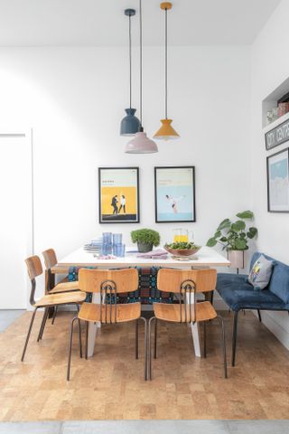 Dining area with white plywood table, an assortment of chairs, three multicolour pendant lights overhead and framed prints of famous films