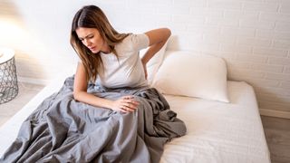 A woman with long dark hair holds her back which is sore after sleeping on a mattress that's too firm for her