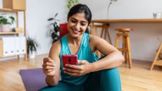 Woman using the best workout apps on phone screen, sitting on yoga mat in the middle of living room at home smiling