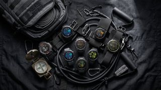 A collage of Garmin smartwatches on a black tarp surrounded by climbing ropes, a compass, and a backpack.