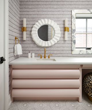 A bathroom with pink cabinets, a white sink and mirror, and textured gray wallpaper