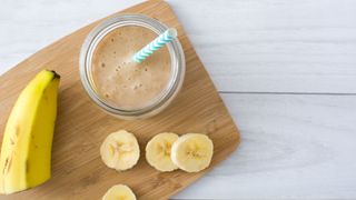 Chopped banana and a banana smoothie in glass with paper straw sitting on wooden chopping board