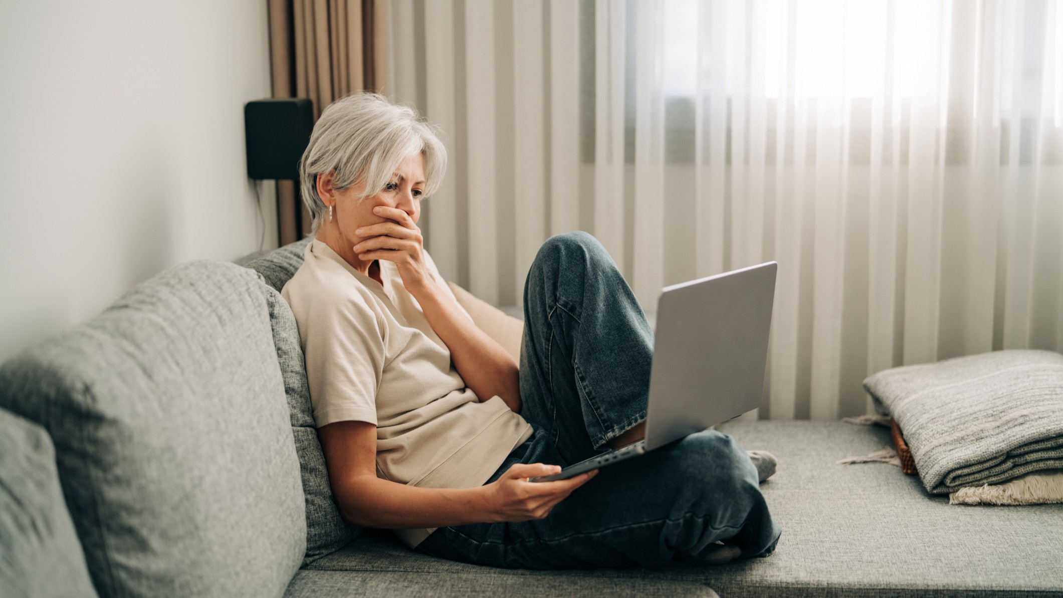 A woman holds her hand over her mouth looking at a laptop while sitting on a comfy couch.