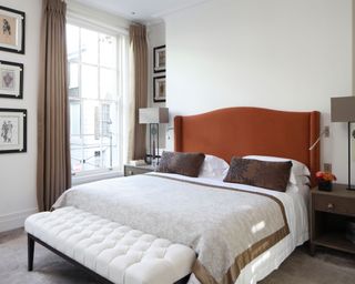 Bedroom with brown upholstered headboard, white bedding and brown cushions and throw, cream ottoman with black frame positioned at the end of the bed, sash window with long brown curtains, brown carpet, dark wooden bedside tables with black and brown table lamps