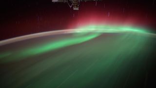 a wash of green aurora, crowned in red, wisps over the top of a night-shadowed earth.