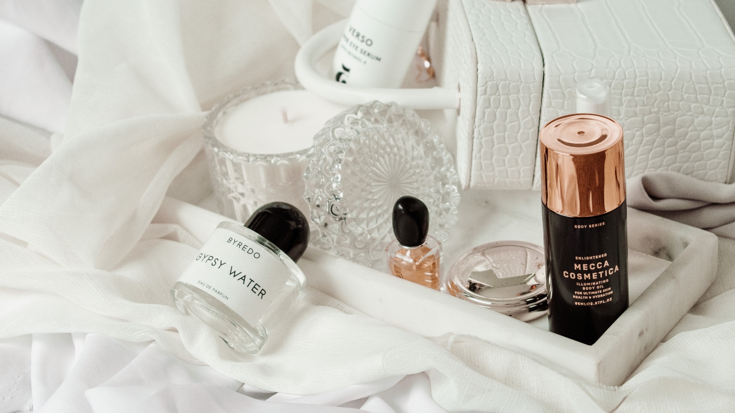 White sheets with a white handbag and white marble tray holding different perfumes and candles