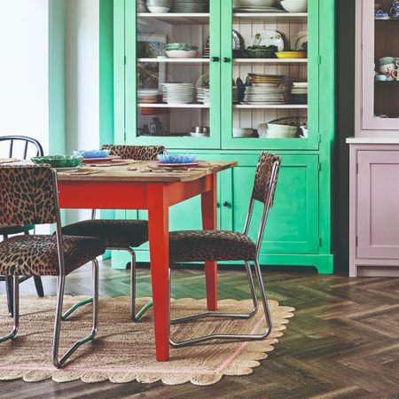 A dining room with green and pink-painted display cabinets, a red table, leopard-print chairs and scalloped jute rug