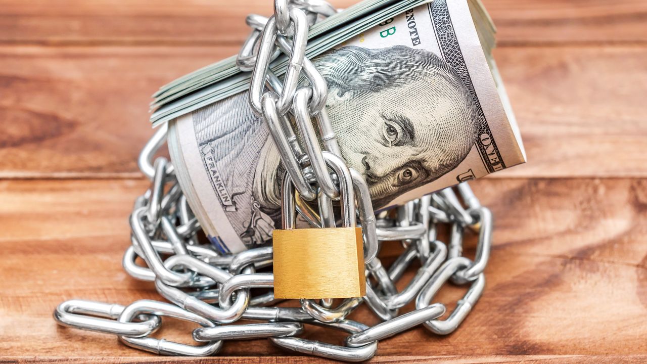 A roll of cash is bound by a chain that&#039;s secured with a padlock.